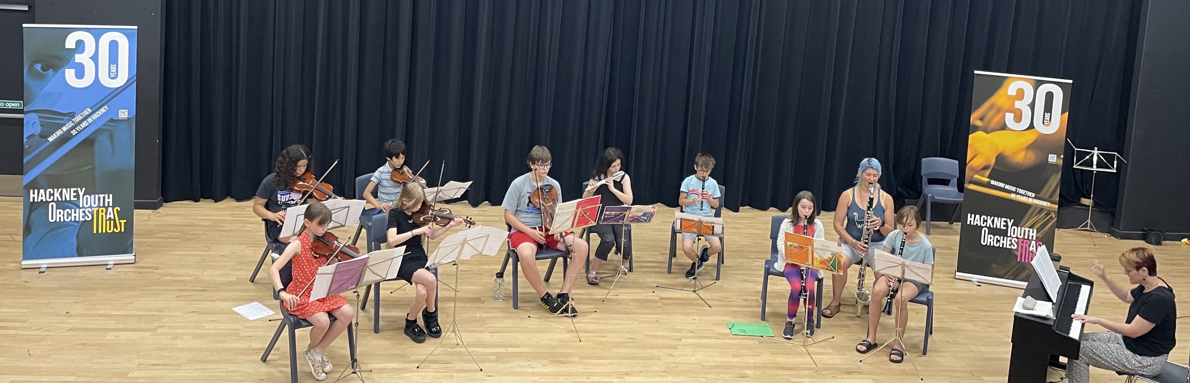 HYOT's Junior Orchestra performing on stage at Stoke Newington School's Theatre. On either side of them there are banners celebrating HYOT's 30th anniversary.